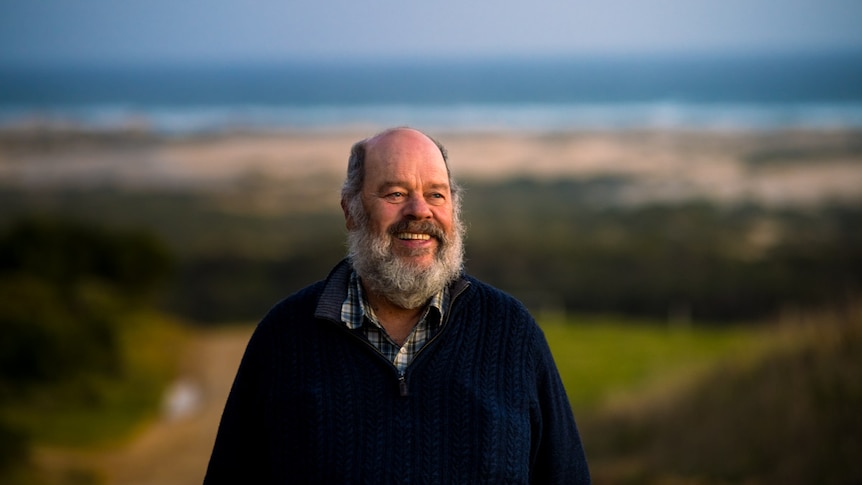 A balding Aboriginal man with a white beard smiles with his green landscape and the sea in the background