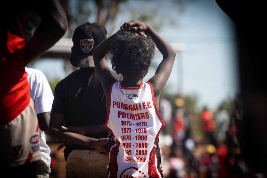 A young boy wearing a red and white football jersey faces away and holds his hands on his head