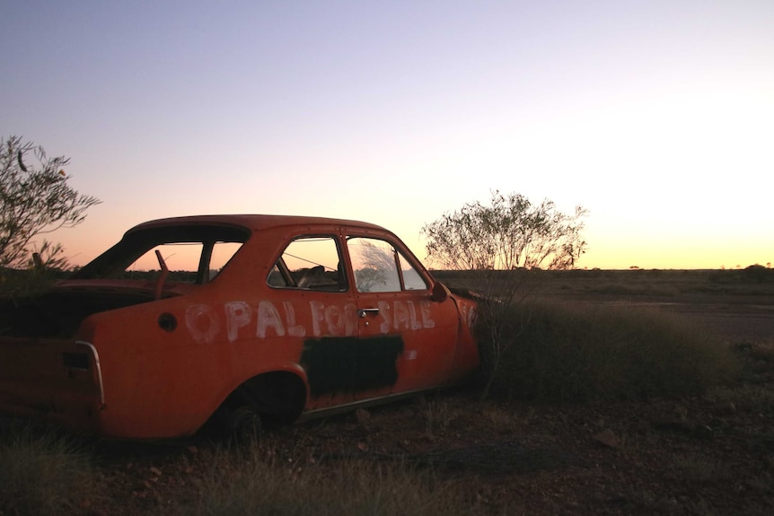 An old dilapidated car sits on blocks on the side of a road. "Opals for Sale" is painted on the side.