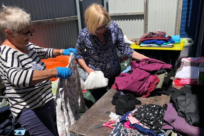 Two middle-aged women sort through piles of coloured clothing on a bench, with corrugated iron walls behind them.