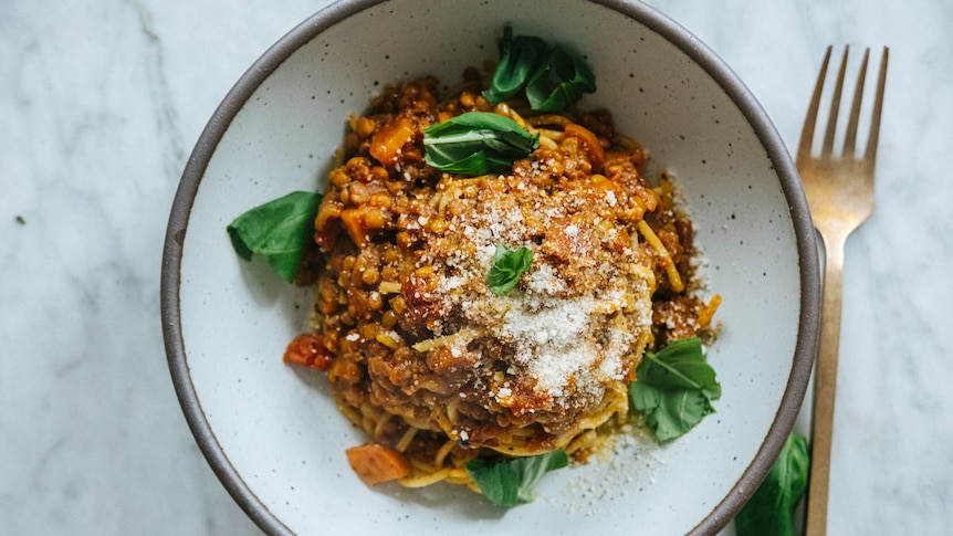 A speckled white bowl of lentil bolognese ragu topped with vegan parmesan cheese and fresh basil.