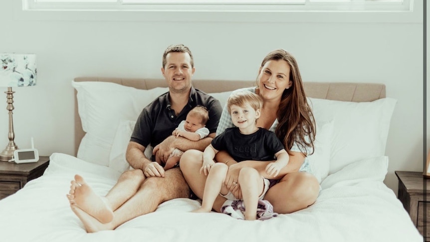 Anthea Marsh sits on a bed with her family smiling.