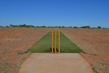 A fake turf cricket pitch and cricket stumps on a rocky desert oval under a clear blue sky.