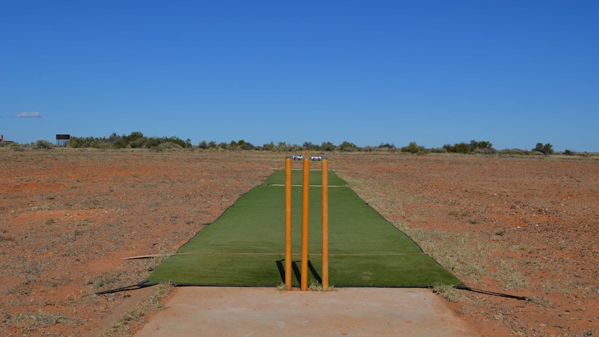 A fake turf cricket pitch and cricket stumps on a rocky desert oval under a clear blue sky.