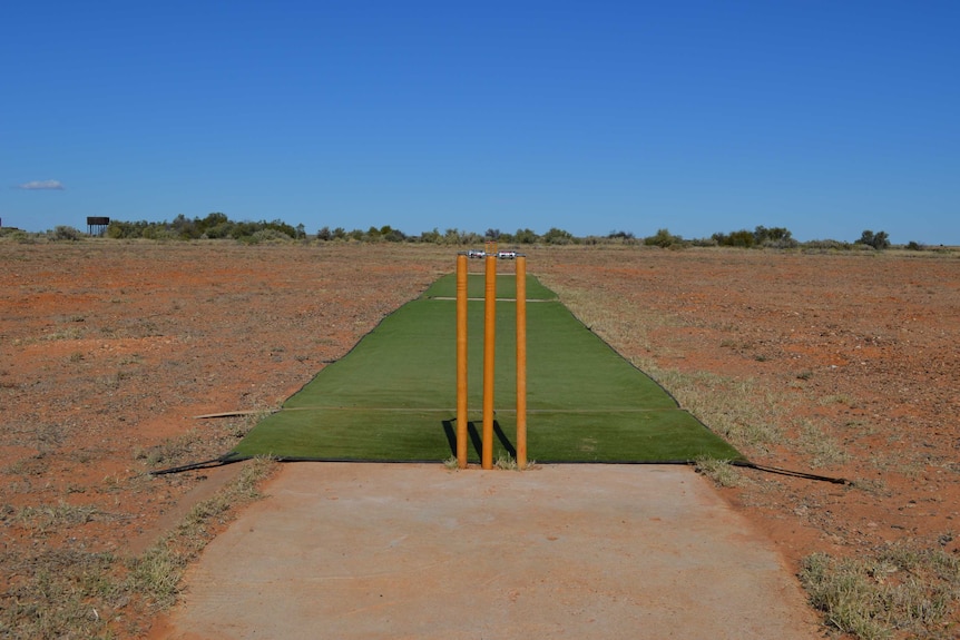 A fake turf cricket pitch and cricket stumps on a rocky desert oval under a clear blue sky.