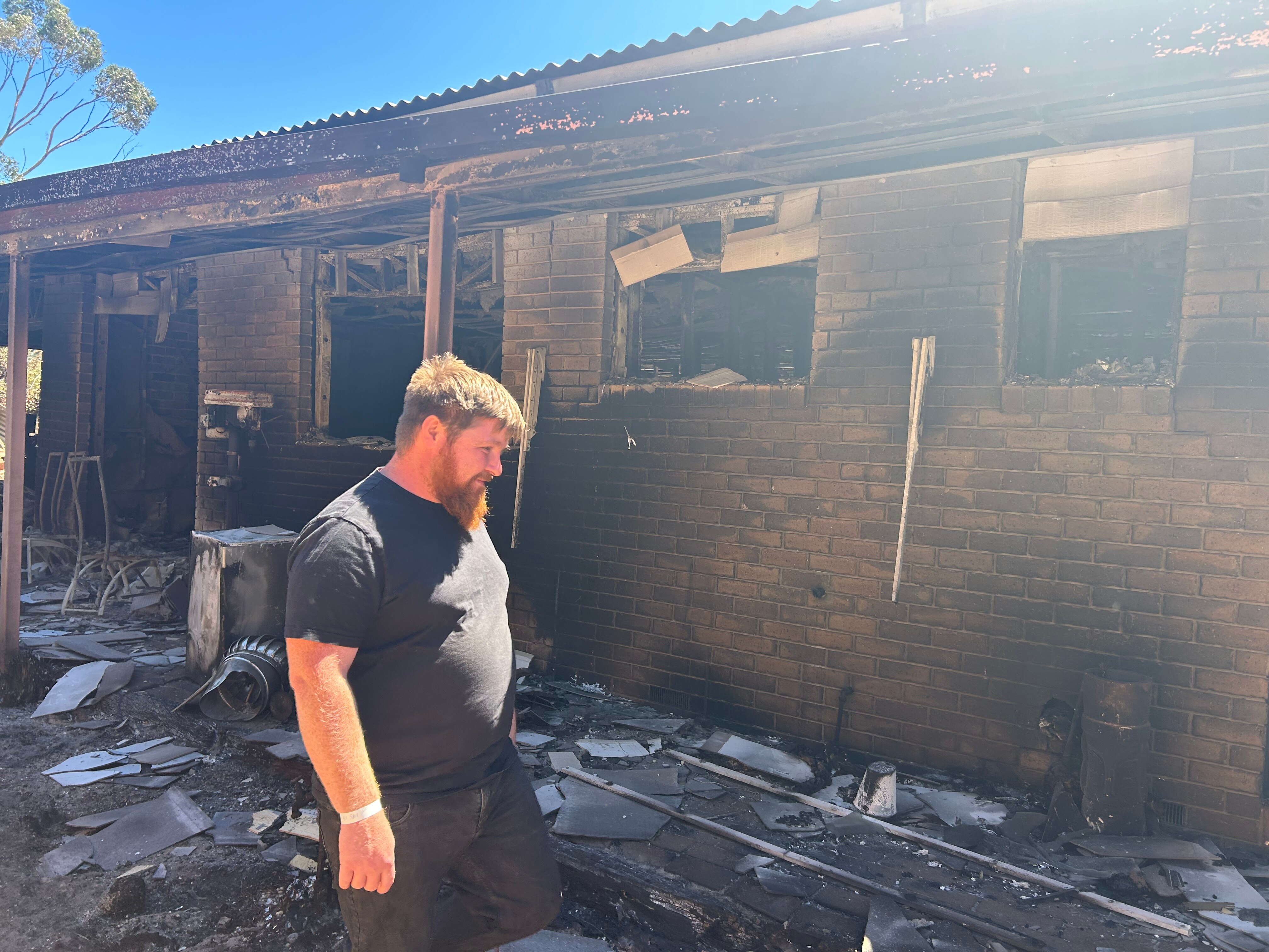 Damon Hendricksen wears a black tshirt and shorts and stands among the burnt debris of his blackened brick home.