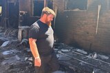 Damon Hendricksen wears a black tshirt and shorts and stands among the burnt debris of his blackened brick home.