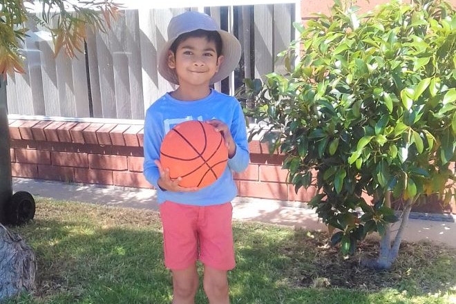 A five-year-old boy stands in a garden in front of a red brick house, holding a basketball in both hands.