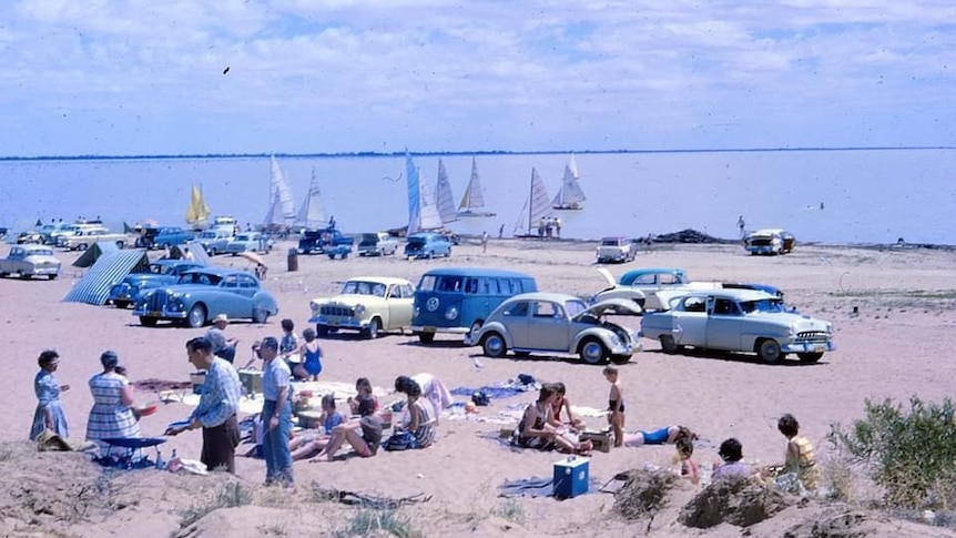 Families gathering on sand near water with cars and boats.