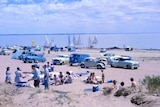 Families gathering on sand near water with cars and boats.