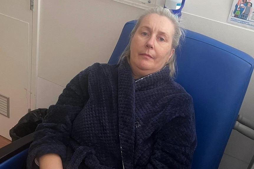 A woman in a dressing gown sits on a hospital chair.