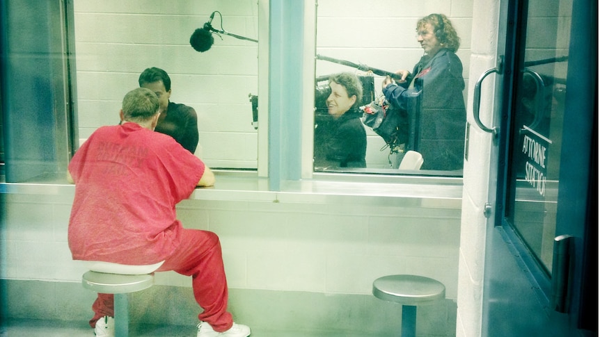 Blue-washed photo of a prisoner talking to a visitor through a glass booth as a documentary team films