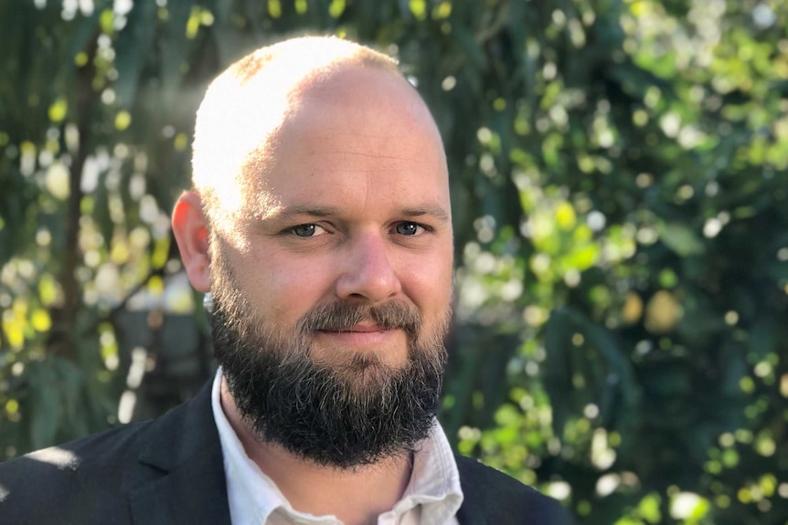 A portrait of Dr Chris Maylea wearing a dark suit jacket and open-necked white shirt.