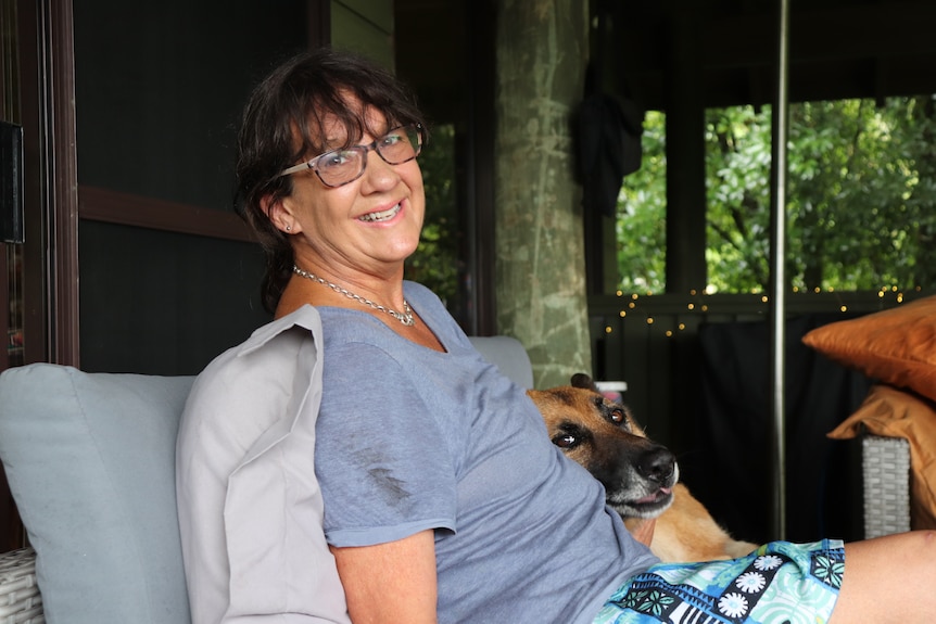 A middle-aged woman with brown hair and glasses sits on a lounge, cuddling her dog.