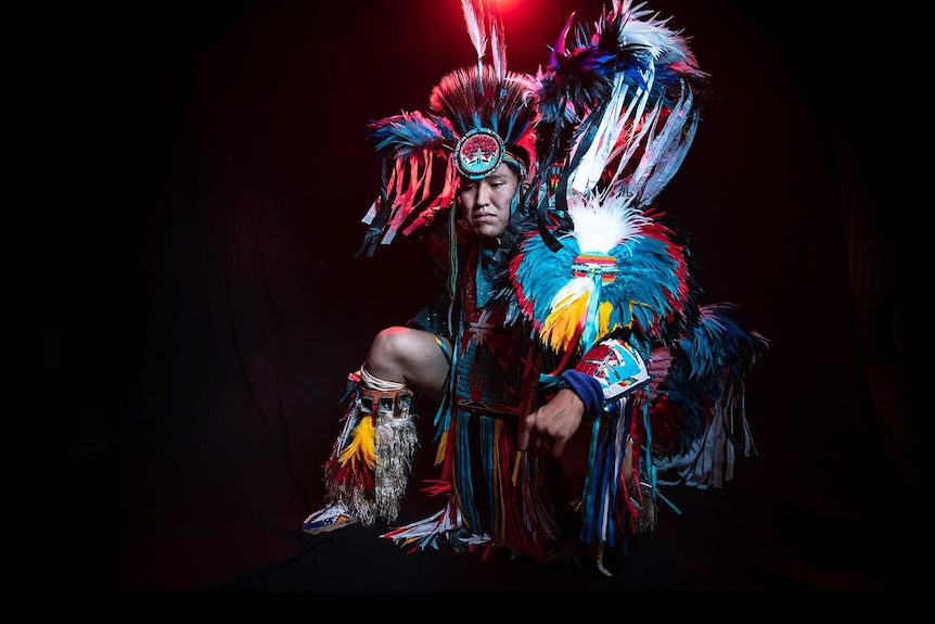 A man in vibrant feathered black, blue and white Native American regalia dances in front of black background with red light.