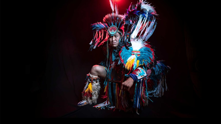 A man in vibrant feathered black, blue and white Native American regalia dances in front of black background with red light.