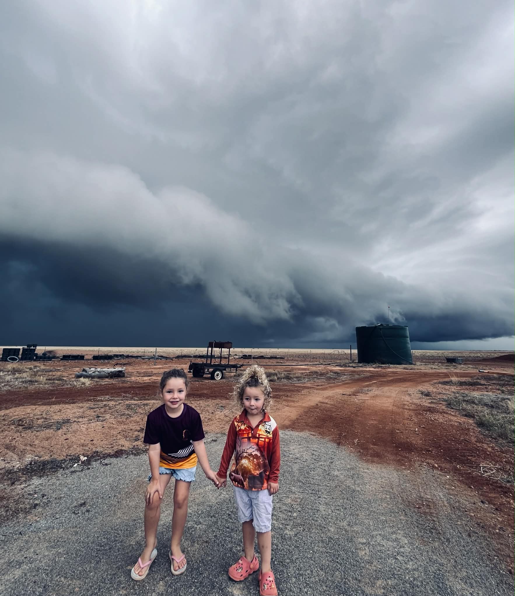Dos niños se encuentran en un camino de tierra con una oscura nube de tormenta detrás de ellos. 