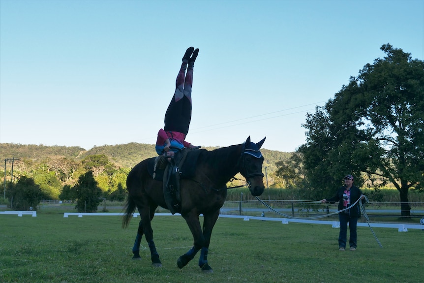 a young woman does a shouldstand on moving horse with coach leading horse off to the side
