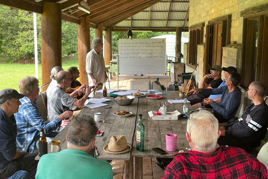 A group of men sitting around a wooden table outside. There's a whiteboard with words.