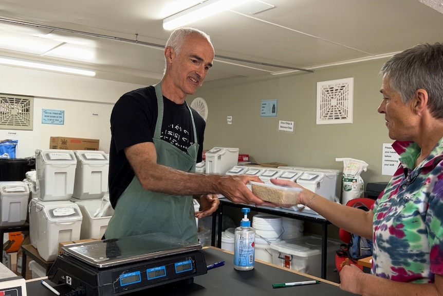 A man with a shaved head stands behind a counter with scales, hands a container to a woman with short grey hair.