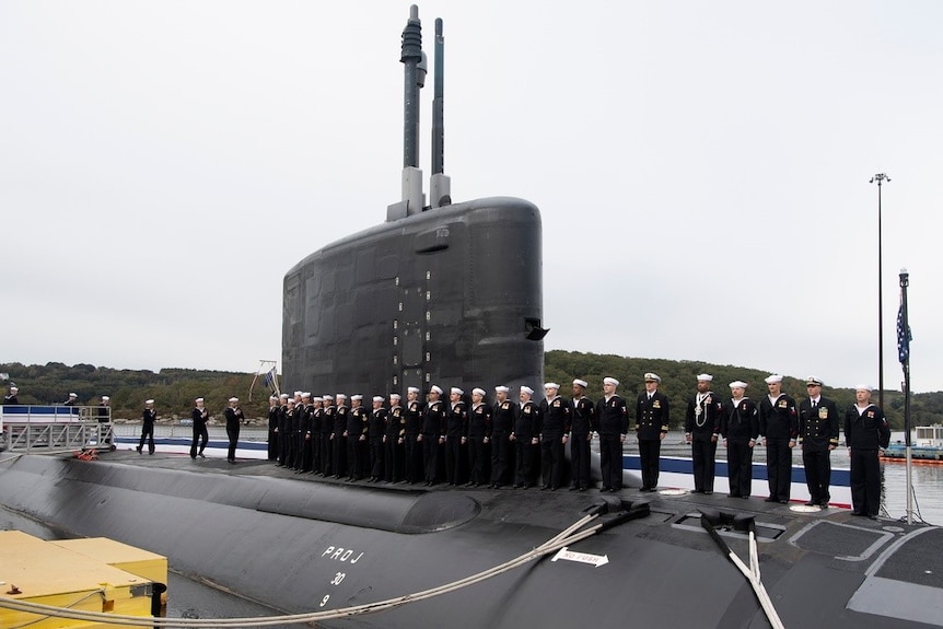 A group of people dressed in Navy uniforms stand on a submarine.