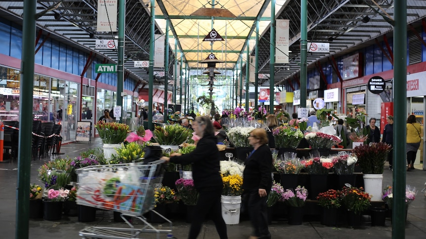Two women walk past a florist under the high, airy frames of the Preston Market.