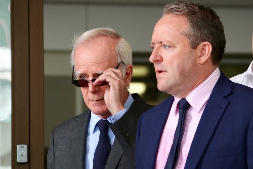 An older man in a suit and sunglasses walks out a building entrance with another man in a suit and pink shirt.