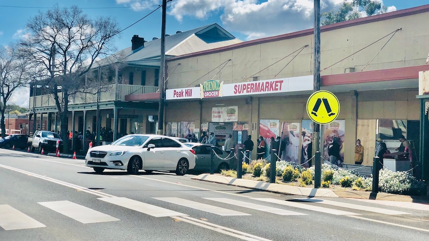 A long line of people wearing face masks extends beyond the supermarket.