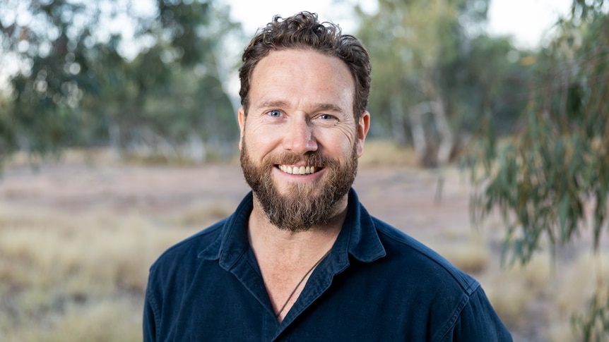 man with brown facial hair and blue shirt smiling at camera in front of blurred bush background