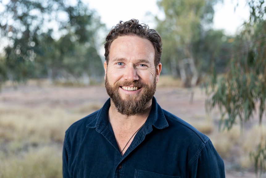 man with brown facial hair and blue shirt smiling at camera in front of blurred bush background