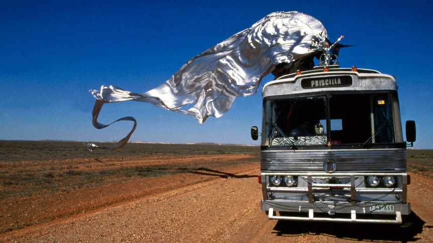 A still from original Priscilla, Queen of the Desert film, with iconic silver wings flying behind a drag queen standing on a bus