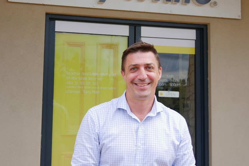 A smiling man with short dark hair, wearing a business shirt, stands in front of real estate agency.