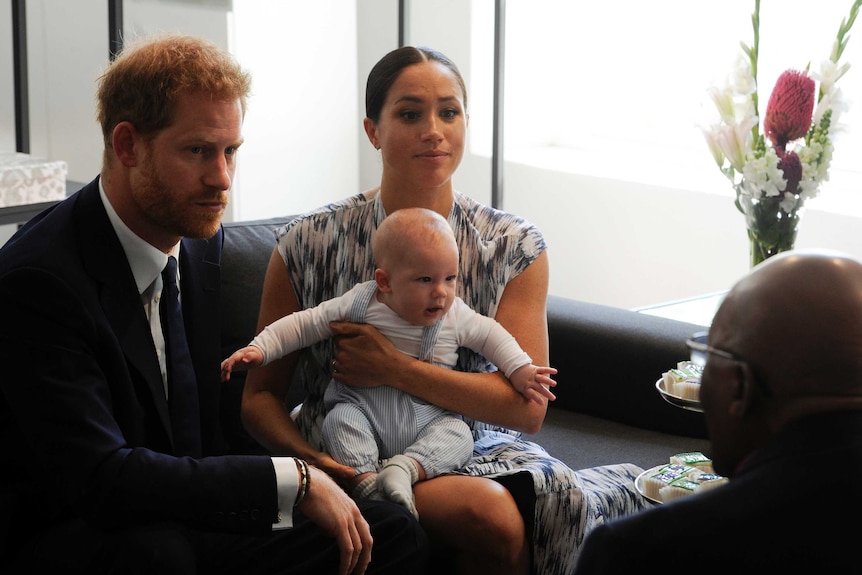 A baby sits on Meghan's lap. Harry sits to her right. They are speaking to Desmond Tutu, whose back is to camera.