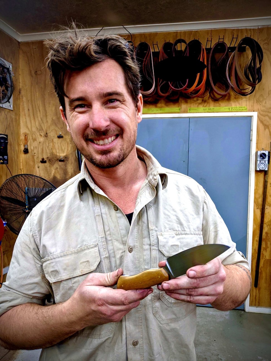 A young man holds up a knife he's made, smiling.