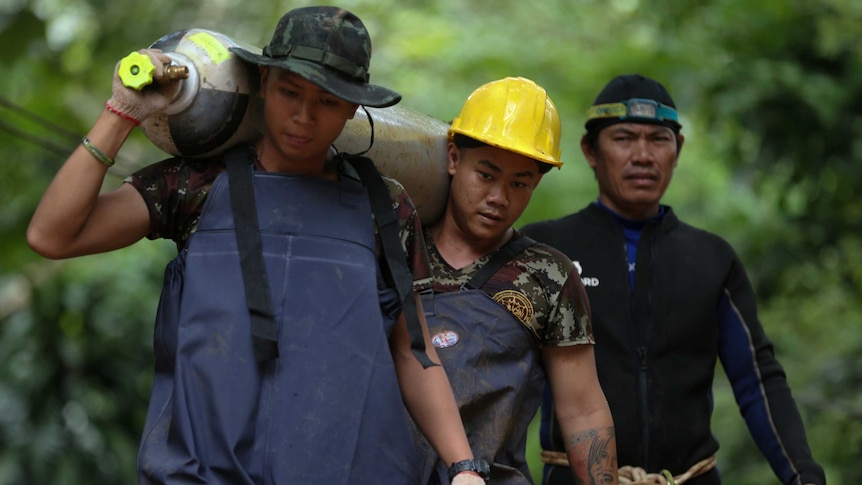 Two workers, one wearing an army hat and one a yellow hard hat, carry an oxygen tank with a diver walking behind them