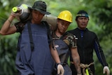 Two workers, one wearing an army hat and one a yellow hard hat, carry an oxygen tank with a diver walking behind them