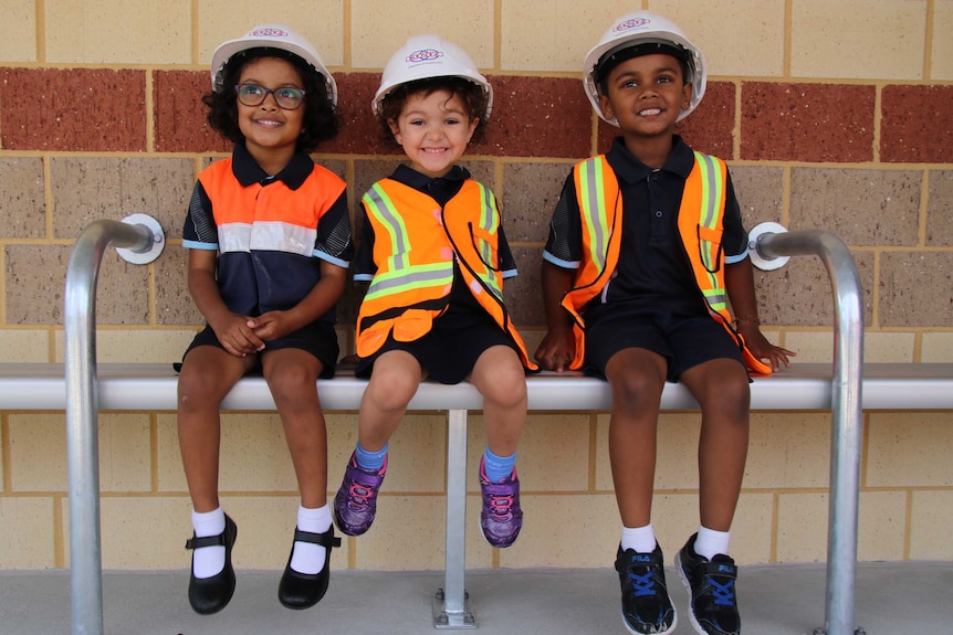 Three small children wear safety helmets and high-vis vests. They have big smalls and are adorable.
