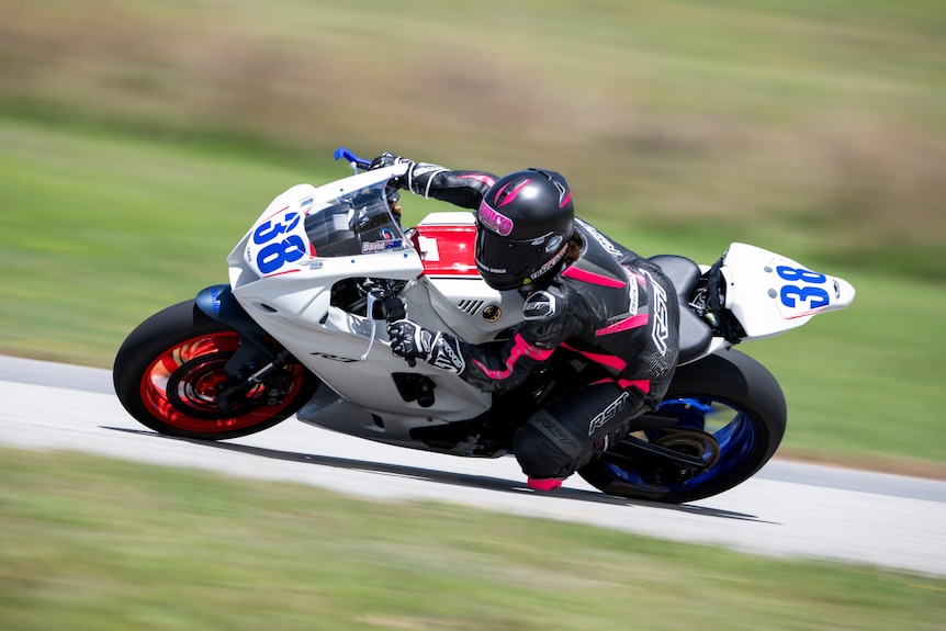 A person wearing black and pink protective gear rides a white, blue, and black motorbike at high speed on a race track.