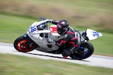 A person wearing black and pink protective gear rides a white, blue, and black motorbike at high speed on a race track.