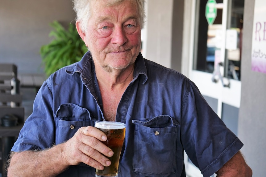 An older man with grey hair, blue eyes and a blue shirt sits outside with a beer in hand.