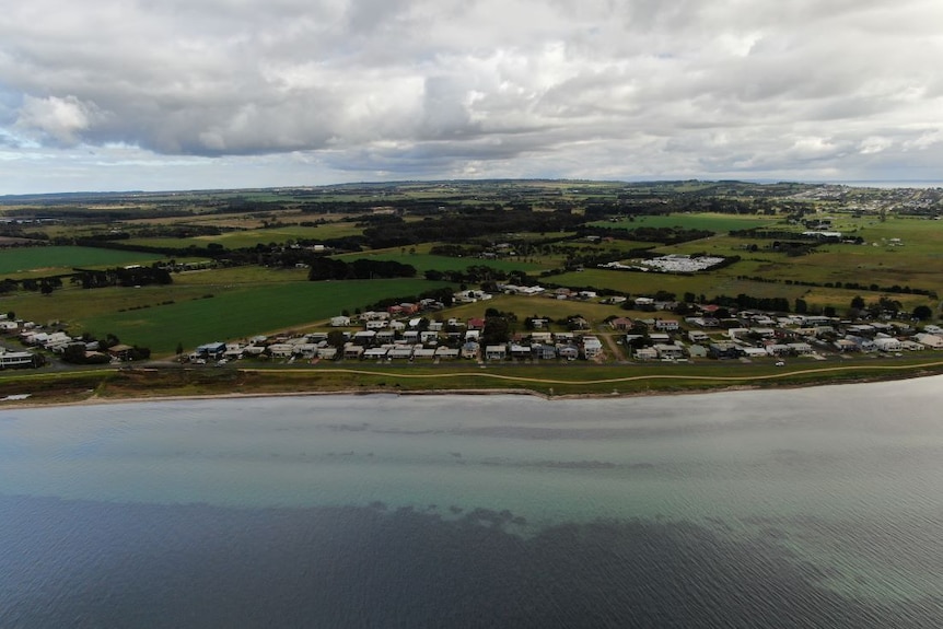 A photograph taken by a drone shows a small cluster of homes beside a bay, with green farmland in the background.