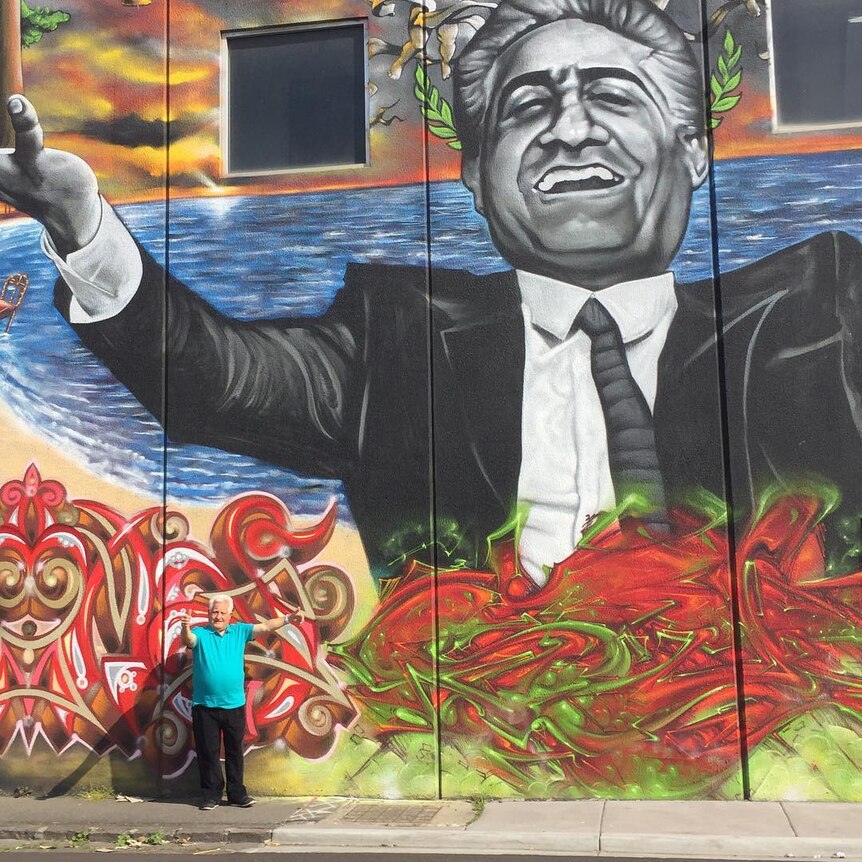 Franco Cozzo standing beneath a giant colourful artistic mural of himself on the side of a building.