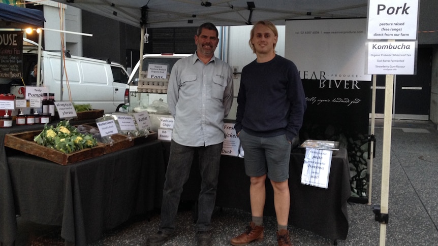 A farmer and worker stand in front of their stall at the farmers' markets.