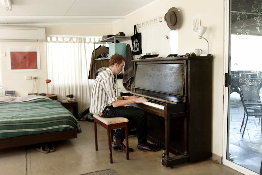 Tall man playing upright piano in bedsit. Bed is made. 4WD and chairs visible through sliding door.
