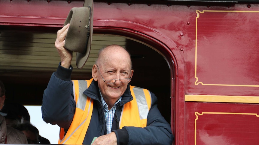 a man stands in a train, looking out of a window and holding his hat up