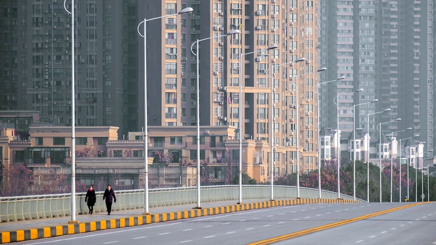 You view an empty six-lane bridge apart from two pedestrians with high rise buildings rising up behind them.