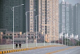 You view an empty six-lane bridge apart from two pedestrians with high rise buildings rising up behind them.