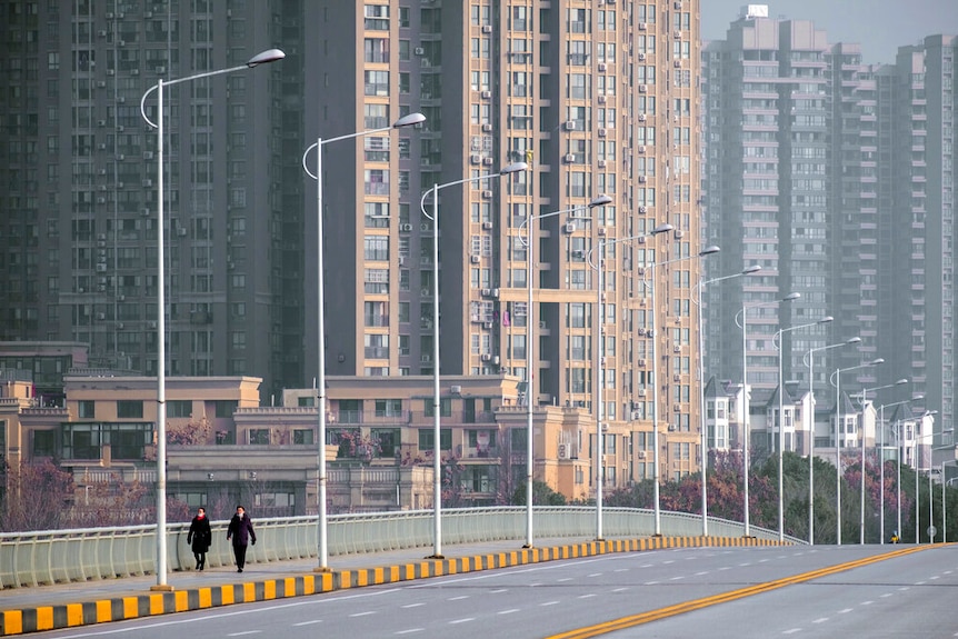 You view an empty six-lane bridge apart from two pedestrians with high rise buildings rising up behind them.