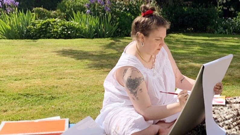Lena Dunham sits in a Welsh garden painting while wearing a white dress.