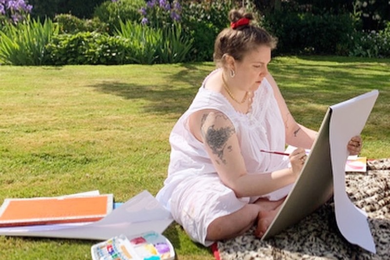 Lena Dunham sits in a Welsh garden painting while wearing a white dress.